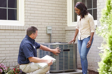 American Home Shield technician inspecting an outdoor air conditioning unit while a homeowner looks on outside a brick house