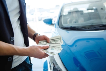 Person counting hundred-dollar bills next to a blue car after refinancing an auto loan