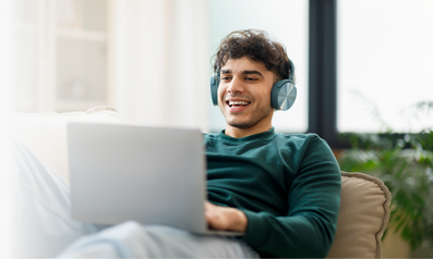 Smiling man wearing headphones using a laptop while relaxing on a couch at home
