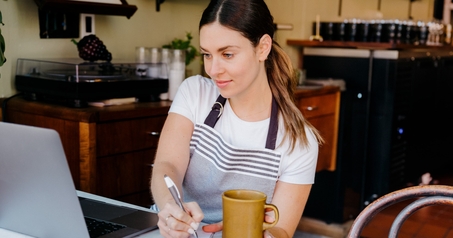 Small business owner in an apron taking notes at a laptop while holding a coffee mug in a cafe