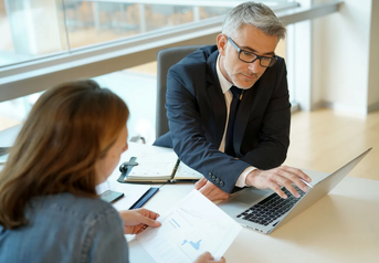 Man in a suit using a laptop across a desk from a woman reviewing a printed chart during a meeting