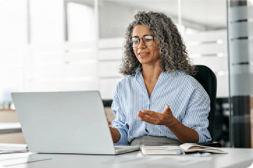 Woman with curly gray hair smiling at a laptop while working at an office desk