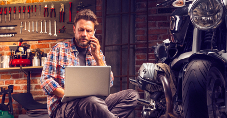 Man on the phone using a laptop next to his motorcycle in a garage workshop.