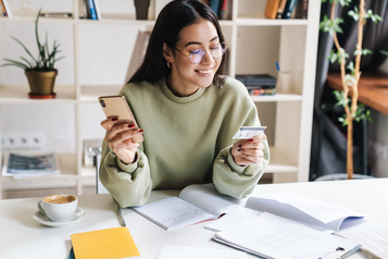 Smiling woman holding a phone and a payment card while doing online banking at her desk.
