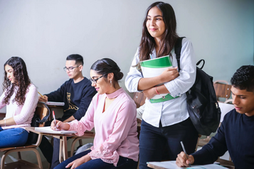 College student standing with a notebook and backpack in a classroom while other students study at their desks