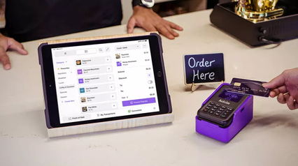Helcim point-of-sale tablet displaying a cafe order with a customer tapping a credit card on a purple Helcim card reader next to an 