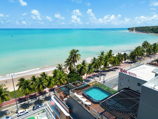 Aerial view of a beachfront hotel with a rooftop pool overlooking a palm-lined avenue and turquoise sea.