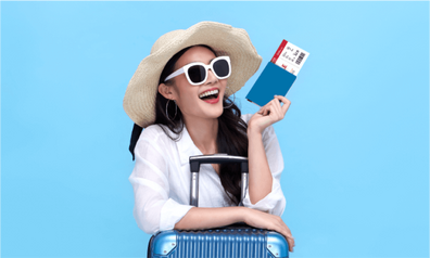 Smiling woman in a sun hat and sunglasses holding her passport and boarding pass while leaning on a suitcase against a blue background