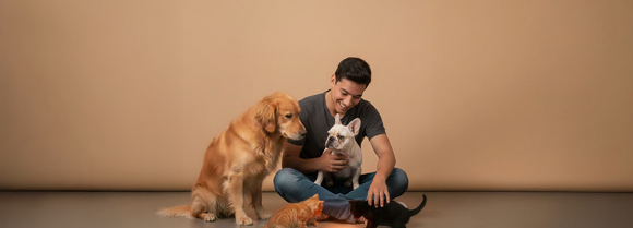 Smiling man sitting cross-legged on the floor with a golden retriever, a French bulldog, and two kittens against a beige backdrop