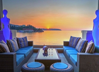 Hotel terrace lounge with rattan sofas and champagne overlooking the sea at sunset.