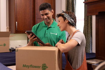 Two Mayflower movers reviewing a moving plan on a tablet beside a packed box in a home