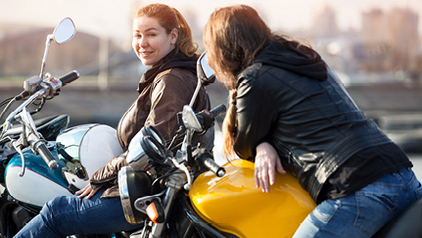Two women in leather jackets sitting on their motorcycles and chatting outdoors.