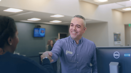 Smiling bank representative shaking hands with a customer across the counter inside a branch.