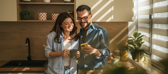 Smiling couple looking at a smartphone together in a sunlit kitchen, one holding a coffee mug