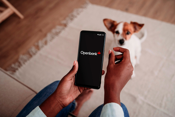 Hand holding a smartphone showing the Openbank app logo on screen with a dog in the background