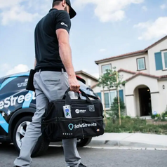 SafeStreets technician carrying a branded equipment bag in front of a suburban home, with a SafeStreets-wrapped vehicle in the driveway.