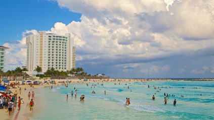 Crowded Cancún beach with swimmers in turquoise water and beachfront resort hotels under a partly cloudy sky.