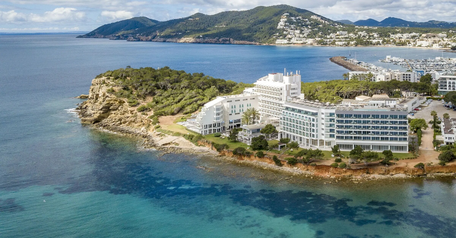 Aerial view of a white oceanfront resort on a rocky peninsula surrounded by turquoise water and coastal hills.