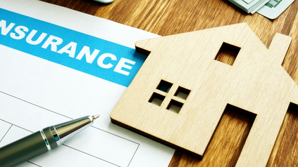 Wooden house cutout resting on a home insurance application form next to a pen on a desk