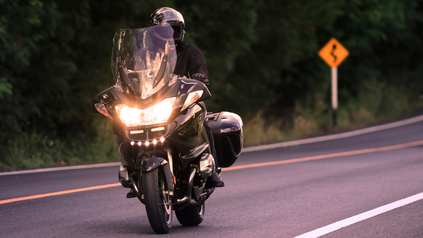 Helmeted rider on a black touring motorcycle with headlight on along a winding tree-lined road.