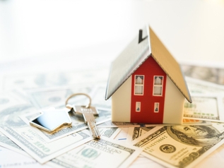Miniature house model and a house-shaped keychain resting on a spread of US hundred-dollar bills